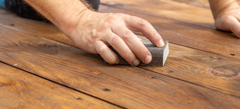 A close-up of a hand sanding a wooden floor with a block sander