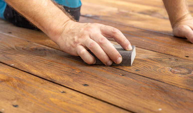 how to sand your own wood floors A close-up of a hand sanding a wooden floor with a block sander